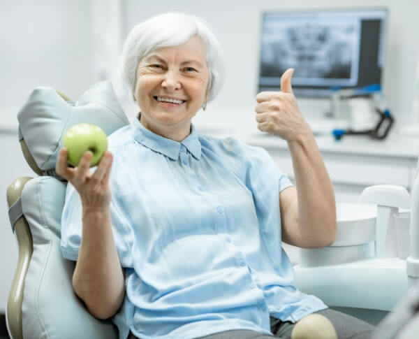 Woman giving thumbs up and holding an apple after dental implant tooth replacement