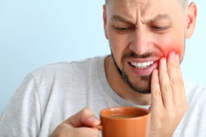 Man in a gray shirt pressing one hand to his inflamed jaw while looking accusingly at his cup of coffee